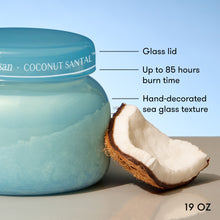 Coconut Santal candle jar with a glass lid and hand-decorated sea glass texture, next to a coconut, on a light blue background.