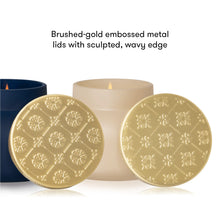 Two round brushed-gold embossed metal candle lid coasters with a sculpted wavy edge, displayed in front of a lit cream candle and a dark blue container on a white background, with text reading 'Brushed-gold embossed metal lids with sculpted, wavy edge'.