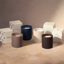 Four matte scented candles in different colors (taupe, navy, cream, and charcoal) arranged in front of their branded white packaging boxes on a textured beige surface.