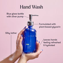 Blue glass hand wash bottle with silver pump held by hands against a pink background
