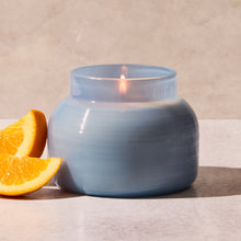 Blue candle in a textured jar with a lit wick, next to an orange slice on a neutral background