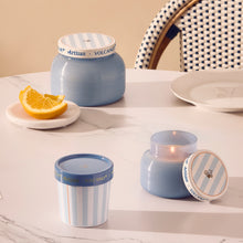 Blue candles with striped lids on a table with a plate of oranges and a chair in the background.