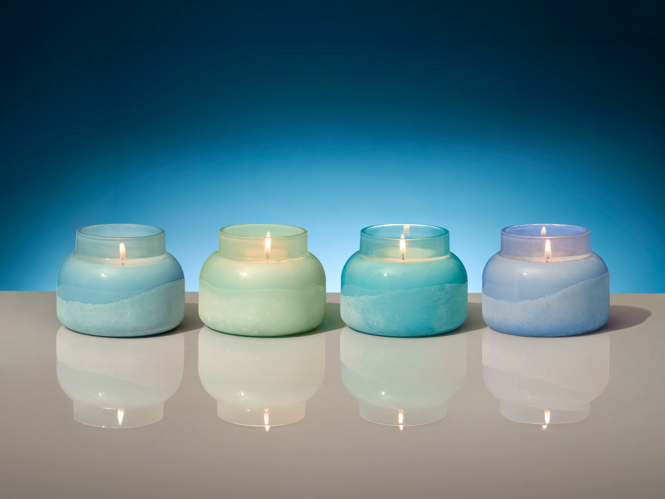 Four lit candles in differently colored glass jars on a reflective surface with a blue gradient background.