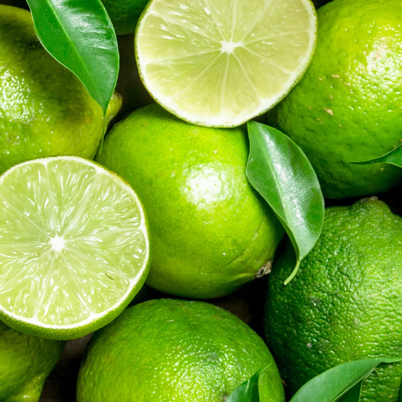 A close-up arrangement of bright green limes, including whole fruits and two halves showing the juicy pulp, with a few glossy green leaves scattered among them.