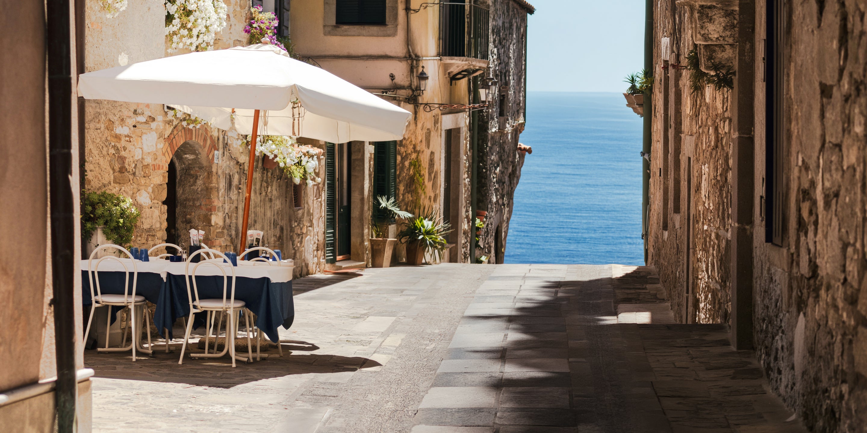 Street view with tables and chairs under an umbrella, leading to a scenic ocean view.