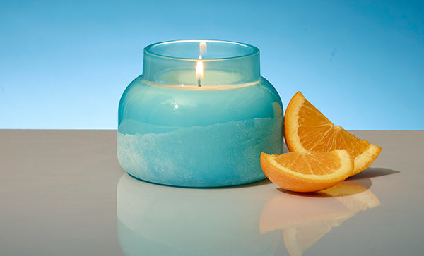 Blue candle with a lit wick and orange slices on a reflective surface with a blue background