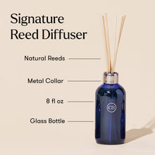 Blue reed diffuser bottle with natural reeds and metal collar on a beige background