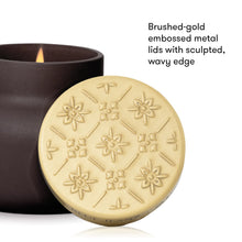 A close-up product shot showing a round brushed-gold metal candle lid with embossed floral and geometric patterns, placed in front of a dark brown candle jar with a visible flame. Text on the right reads, 'Brushed-gold embossed metal lids with sculpted, wavy edge.