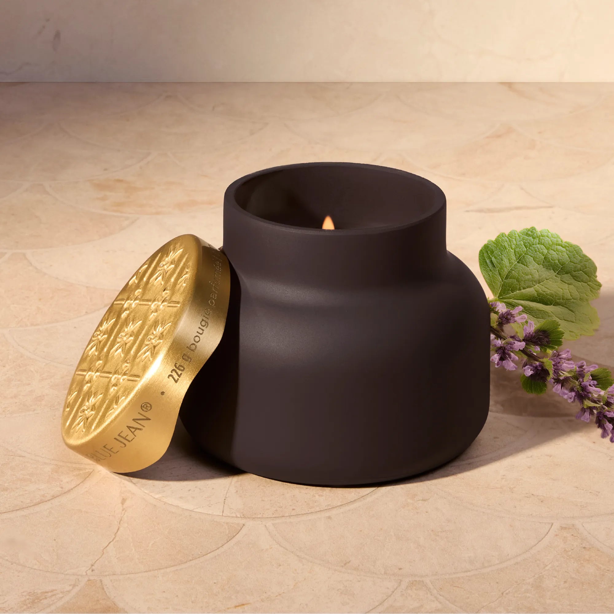 A matte black jar candle with a lit wick sits on a beige stone surface, with its gold-toned lid leaning beside it and a sprig of purple flowers nearby. The lid shows the brand name Jean Paul Gaultier.