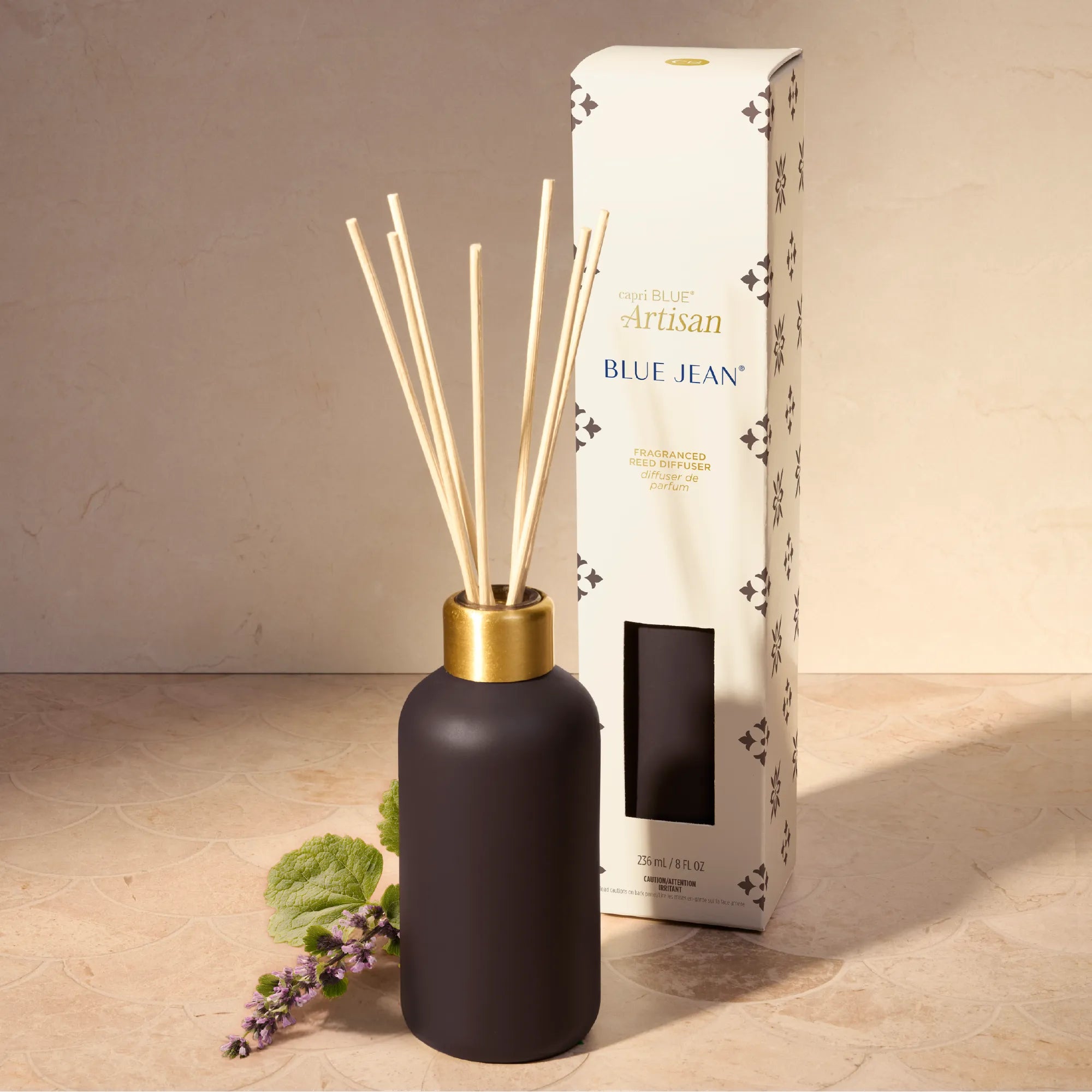 A matte black reed diffuser bottle with a gold neck and natural reeds, shown next to its white branded box on a beige stone surface against a beige wall.