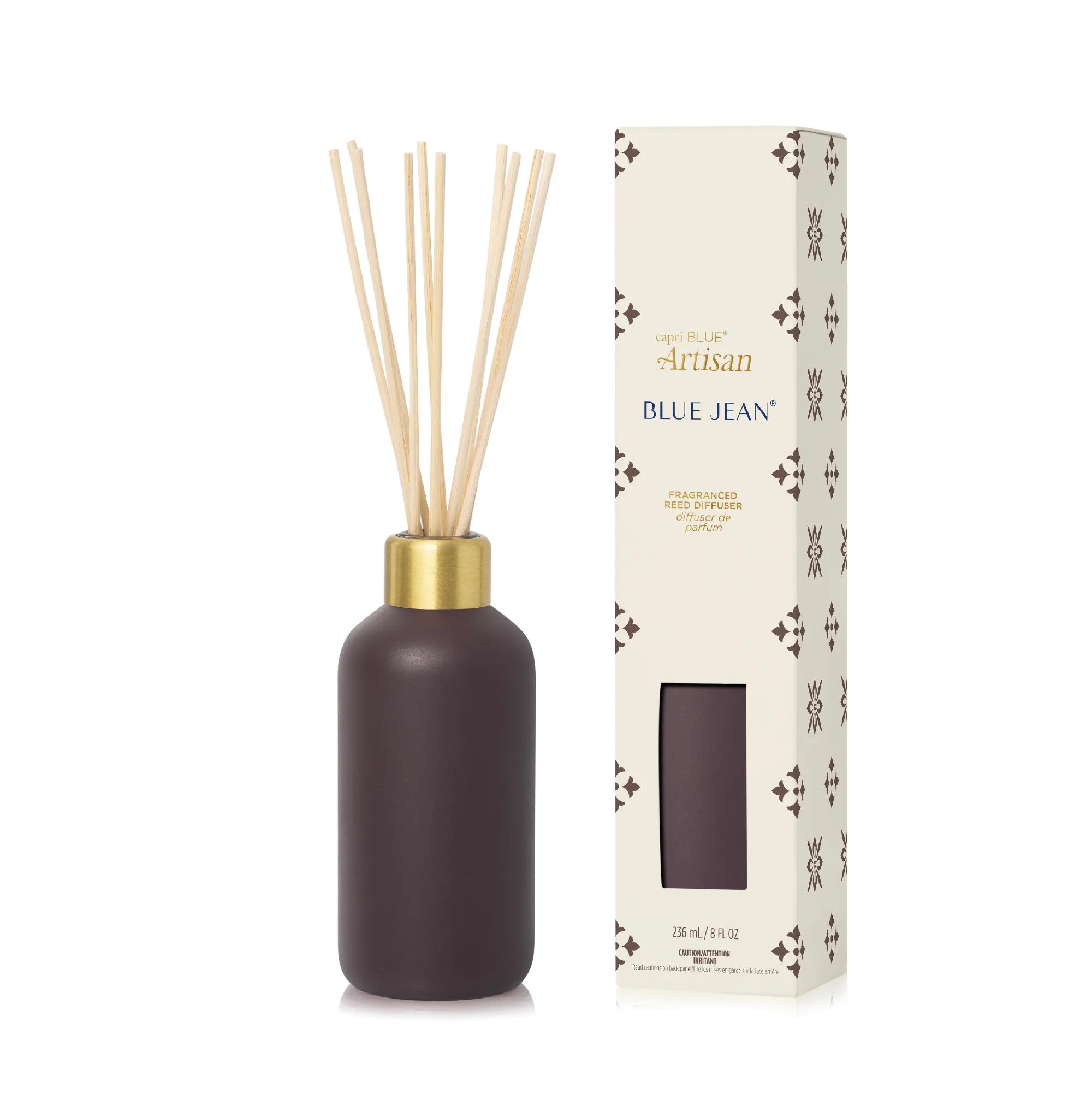 A reed diffuser bottle with natural reeds and a gold collar stands next to its cream-colored packaging box featuring a window and decorative patterns. The bottle is a dark plum-brown color.