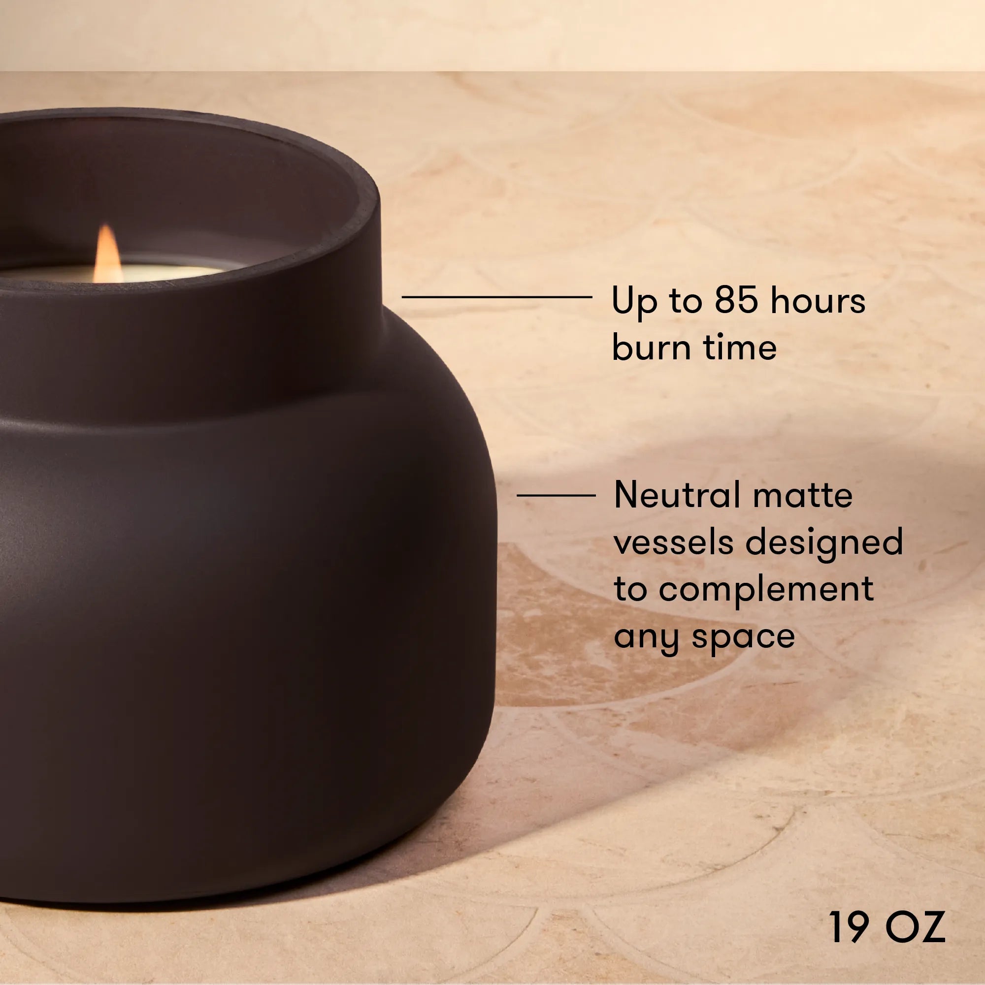 A matte black candle vessel with a lit candle, shown on a beige stone surface with text callouts about 85-hour burn time and neutral matte vessels.