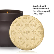 A close-up product shot of a candle tin with its lid leaning in front. The lid is brushed gold with an embossed geometric floral pattern and a wavy edge, while the dark brown candle vessel sits behind it with a small flame visible.