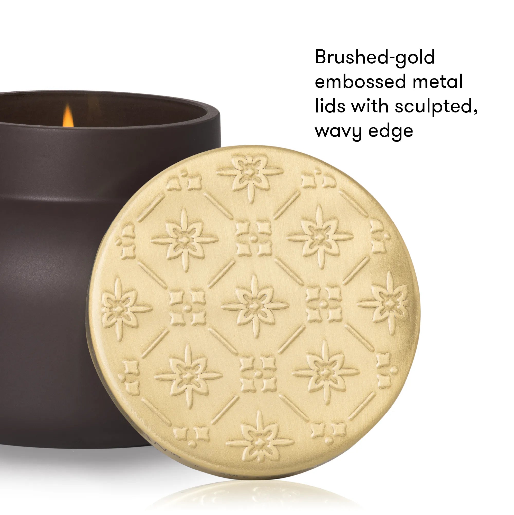 A close-up product shot of a candle tin with its lid leaning in front. The lid is brushed gold with an embossed geometric floral pattern and a wavy edge, while the dark brown candle vessel sits behind it with a small flame visible.
