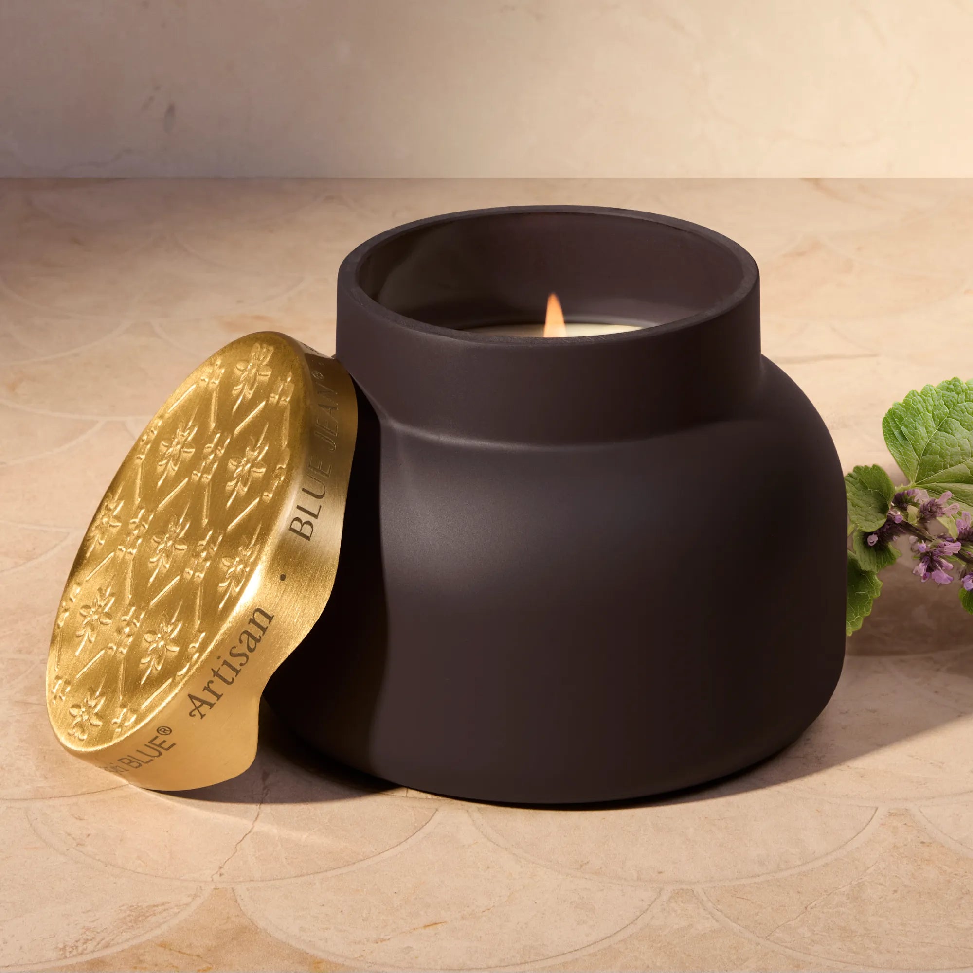 A matte black jar candle with a gold lid leaning beside it on a beige stone surface. The lid is embossed with a floral pattern and the words 'ARTISAN - BLUE JEAN'