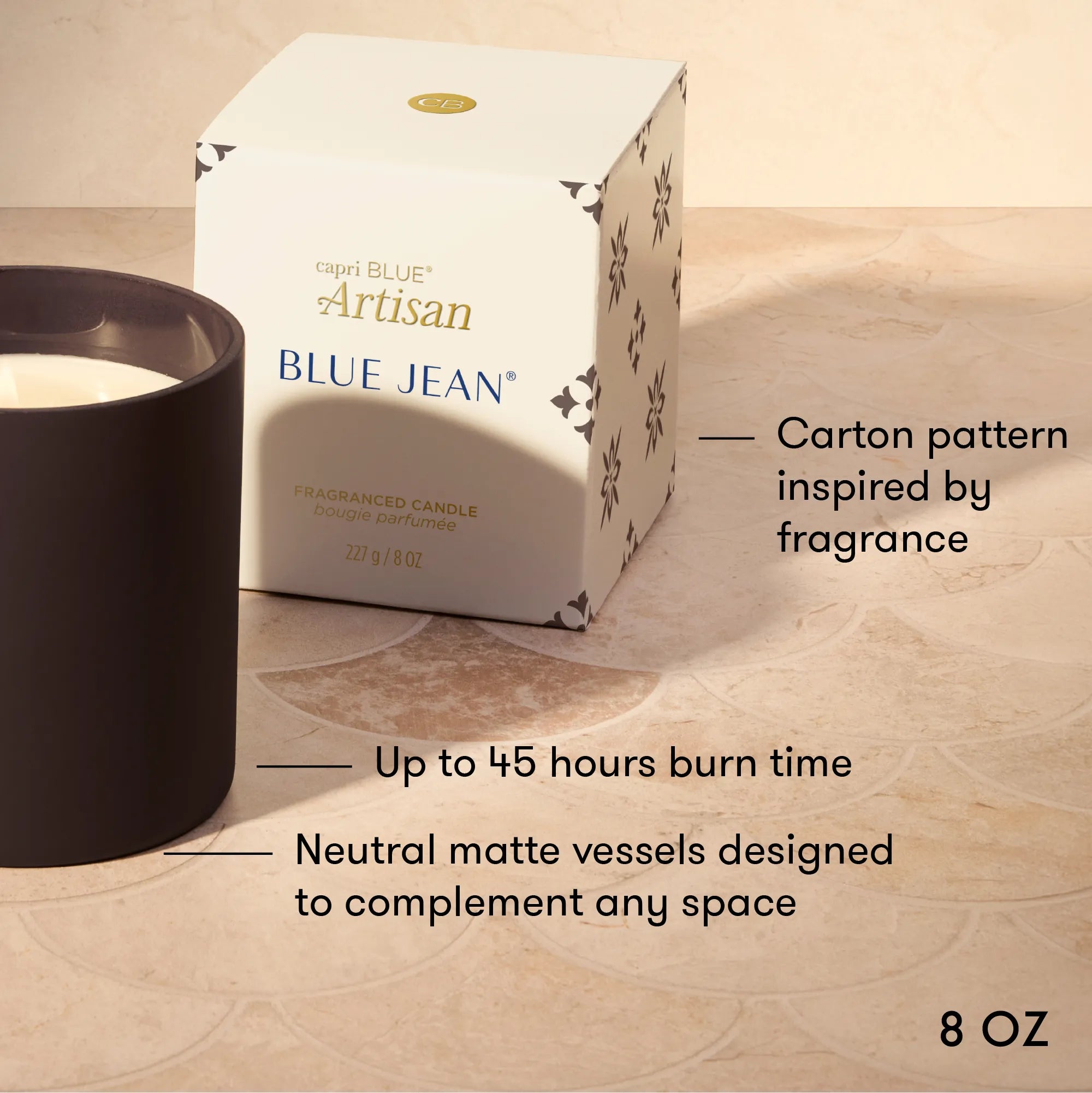 A candle product shown with its branded box on a beige stone-like surface. The candle vessel is matte black and partially visible on the left, with the box labeled 'capri BLUE Artisan BLUE JEAN' and text callouts describing features such as carton pattern inspiration, up to 45 hours burn time, and neutral matte vessels.