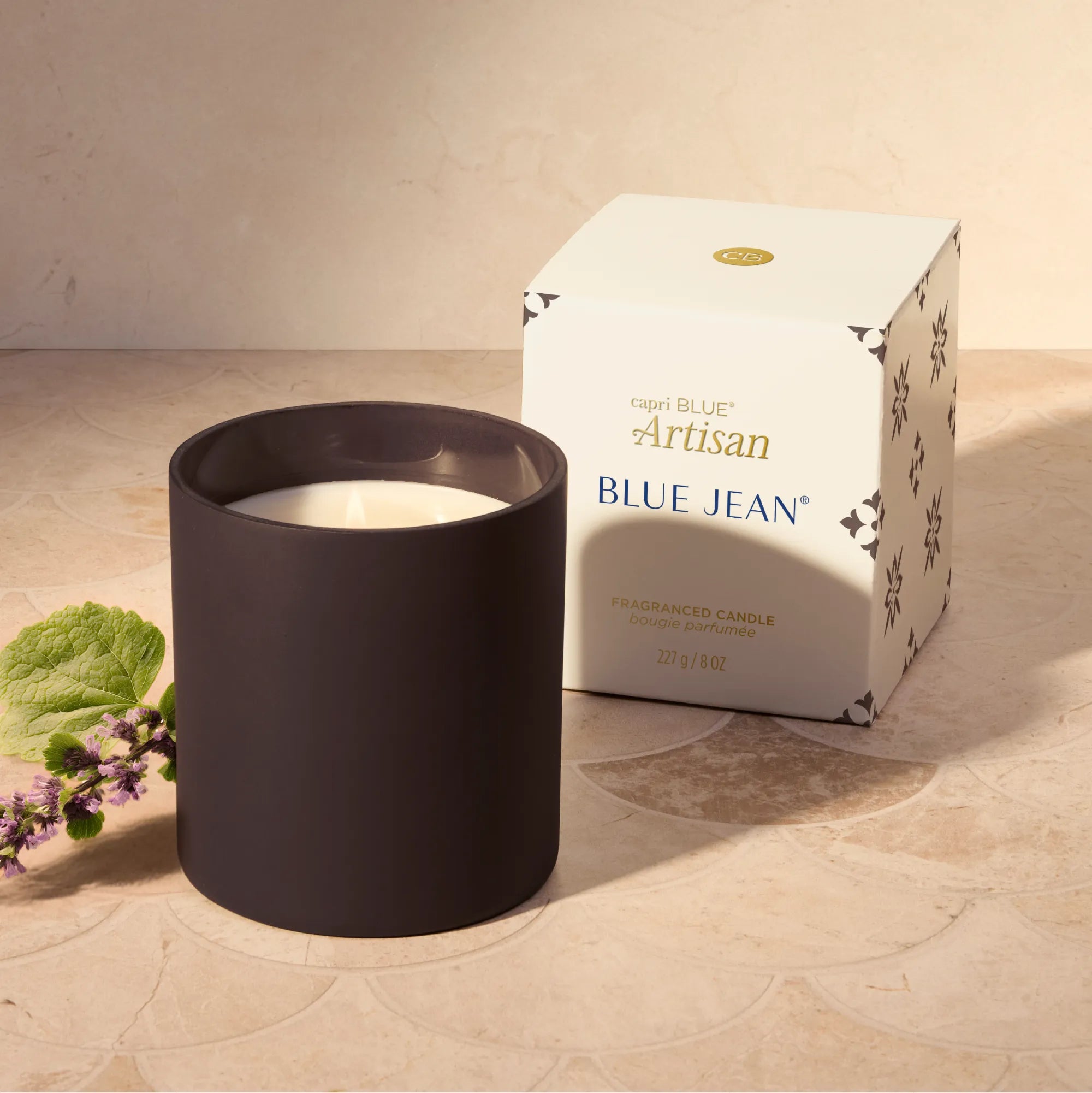 A matte black candle in a cylindrical holder sits next to its white and gold Capri Blue Artisan Blue Jean packaging box on a beige stone surface.
