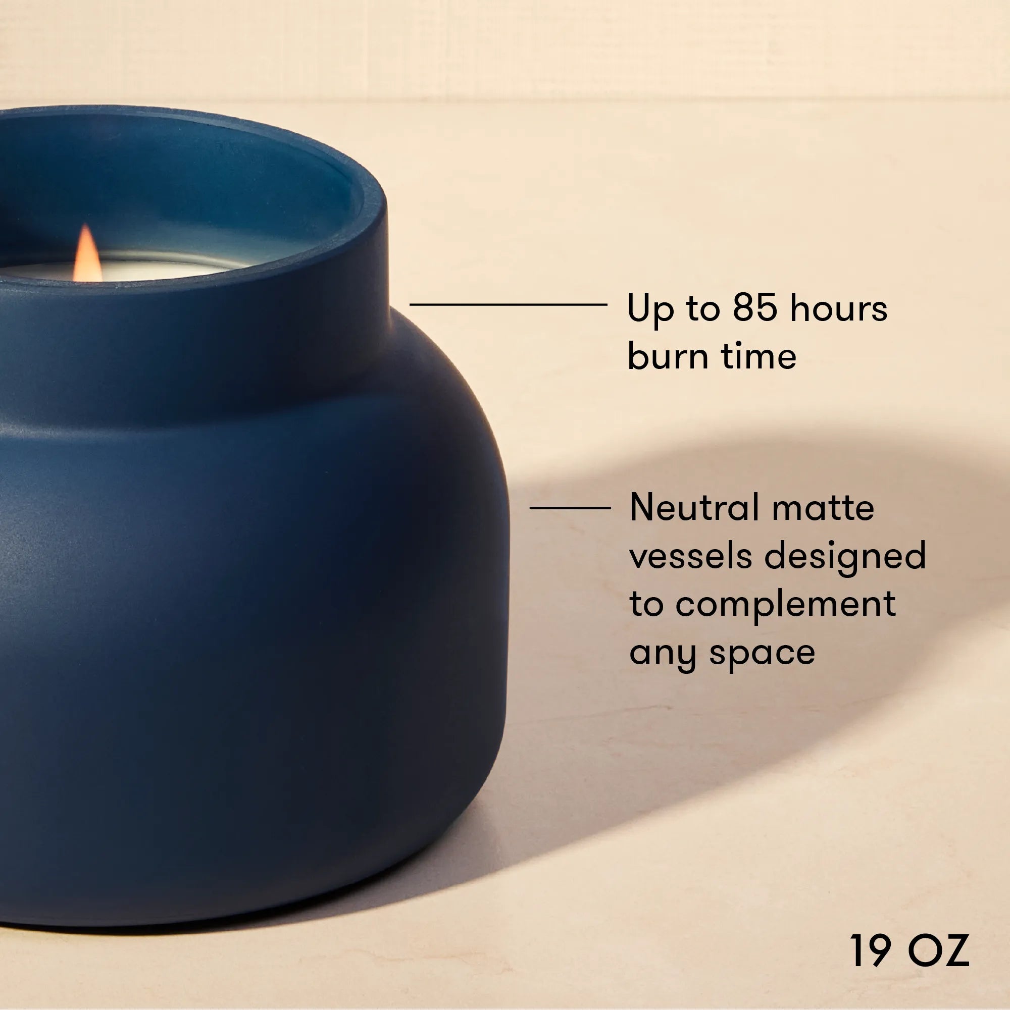 A dark blue matte candle vessel with a lit candle, shown on a warm beige background with text callouts about burn time and neutral matte compatibility. '19 OZ' is printed in the bottom right corner.
