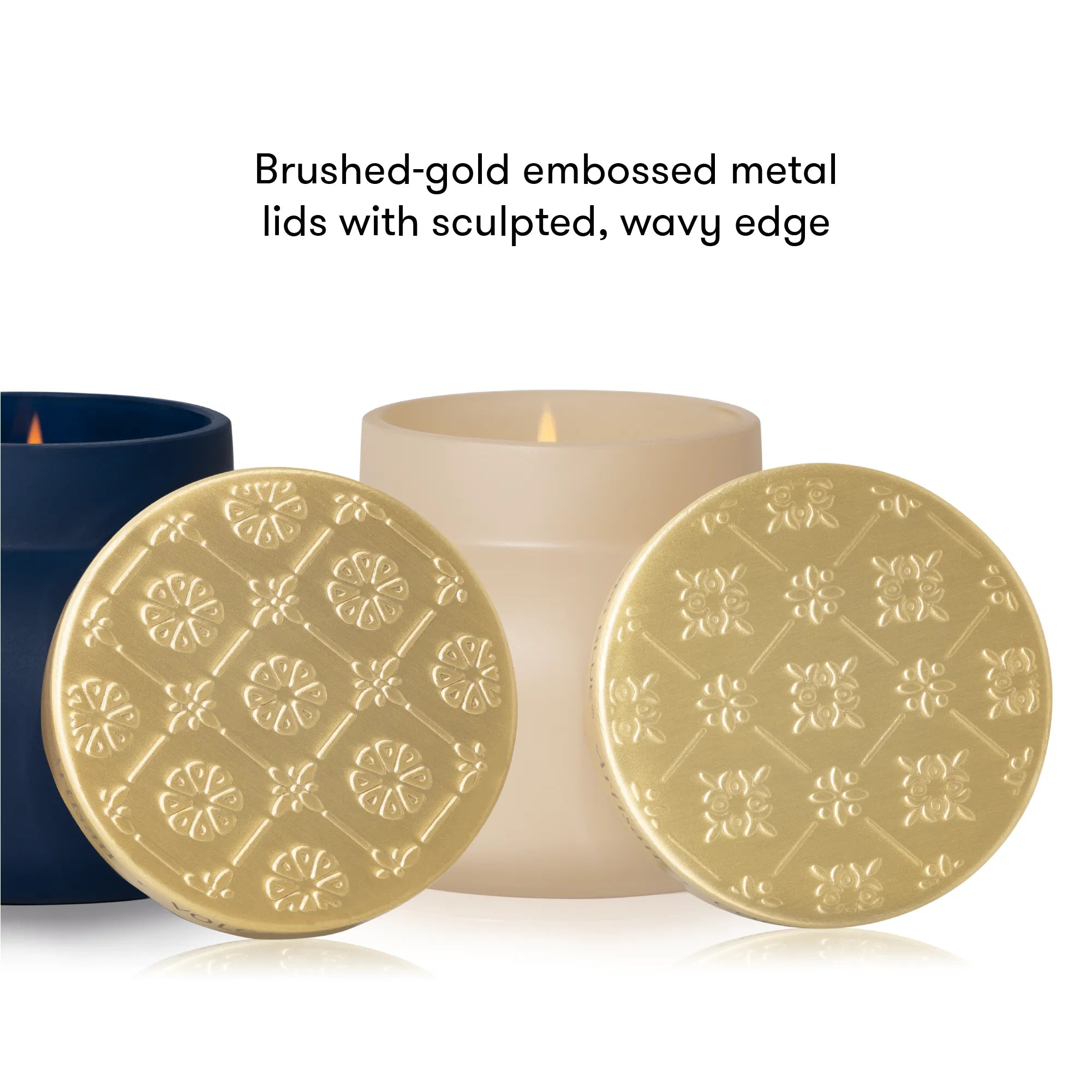 Two round brushed-gold embossed metal candle lid coasters with a sculpted wavy edge, displayed in front of a lit cream candle and a dark blue container on a white background, with text reading 'Brushed-gold embossed metal lids with sculpted, wavy edge'.