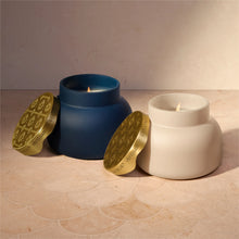 Two ceramic candles in matte blue and off-white pots with gold lids, shown lit on a beige tiled surface against a beige wall.