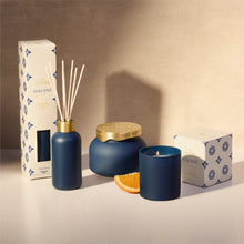 A Volcano scented home fragrance set in matte navy blue, including a reed diffuser bottle with reeds, a jar candle with a gold lid, a candle jar, and branded white packaging boxes, styled with an orange slice on a beige surface.