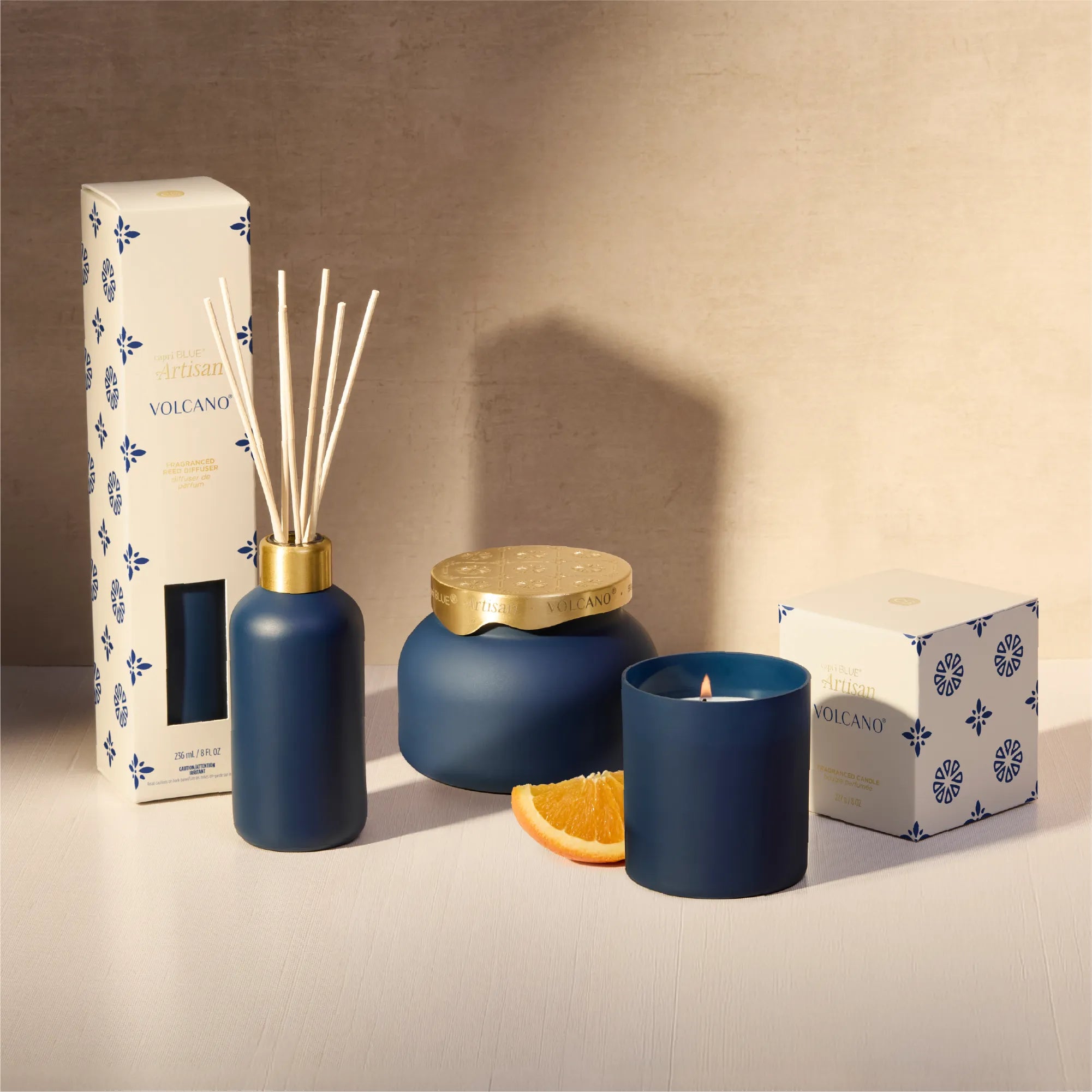 A Volcano scented home fragrance set in matte navy blue, including a reed diffuser bottle with reeds, a jar candle with a gold lid, a candle jar, and branded white packaging boxes, styled with an orange slice on a beige surface.