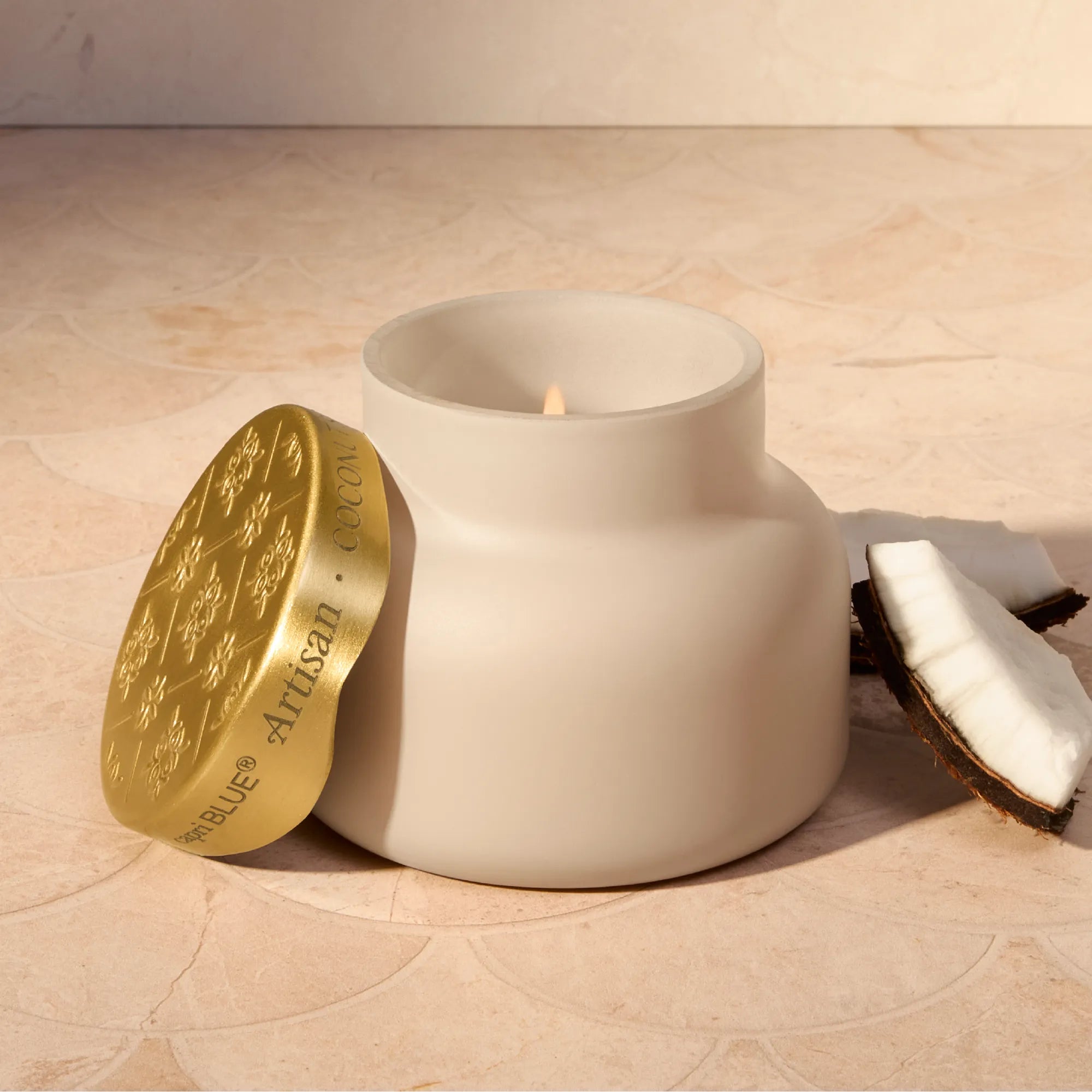 A matte off-white ceramic jar candle with its lid leaning against it on a beige stone surface. The lid is metallic gold with embossed floral patterns and the text 'BLUE ARTISAN'.