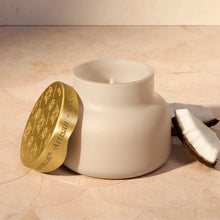 A matte off-white ceramic jar candle with its lid leaning against it on a beige stone surface. The lid is metallic gold with embossed floral patterns and the text 'BLUE ARTISAN'.