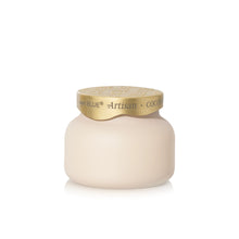 A cream-colored cosmetic jar with a metallic gold lid, featuring the brand name 'capri BLUE' and product name 'Artisan COCONUT' printed on the gold band.