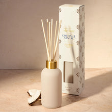 A coconut sandalwood reed diffuser with a matte cream bottle, natural reeds, and a gold collar, shown next to its white packaging box with brown paw print patterns on a beige stone surface.
