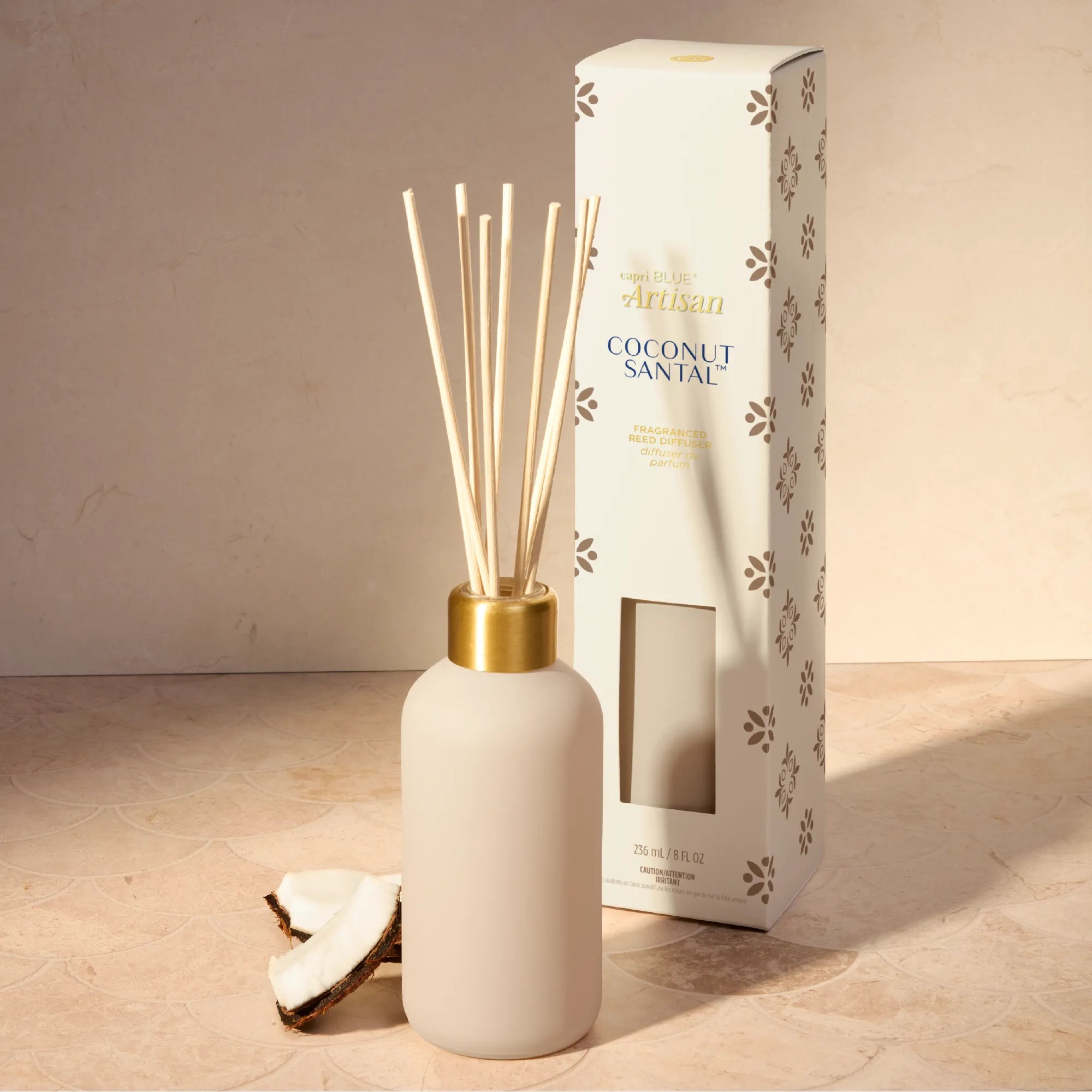 A coconut sandalwood reed diffuser with a matte cream bottle, natural reeds, and a gold collar, shown next to its white packaging box with brown paw print patterns on a beige stone surface.