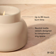 A lit matte ceramic candle holder in a warm off-white/cream tone, shown on a beige stone surface with text callouts reading 'Up to 85 hours burn time' and 'Neutral matte vessels designed to complement any space.'.