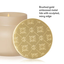 A close-up product shot showing a round brushed-gold metal candle lid with embossed floral patterns and a wavy edge, placed in front of a cream-colored candle. Black text on the right reads, 'Brushed-gold embossed metal lids with sculpted, wavy edge.