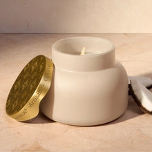 A matte cream-colored ceramic candle jar with a lit wick, shown next to its gold-toned lid on a beige marble surface.