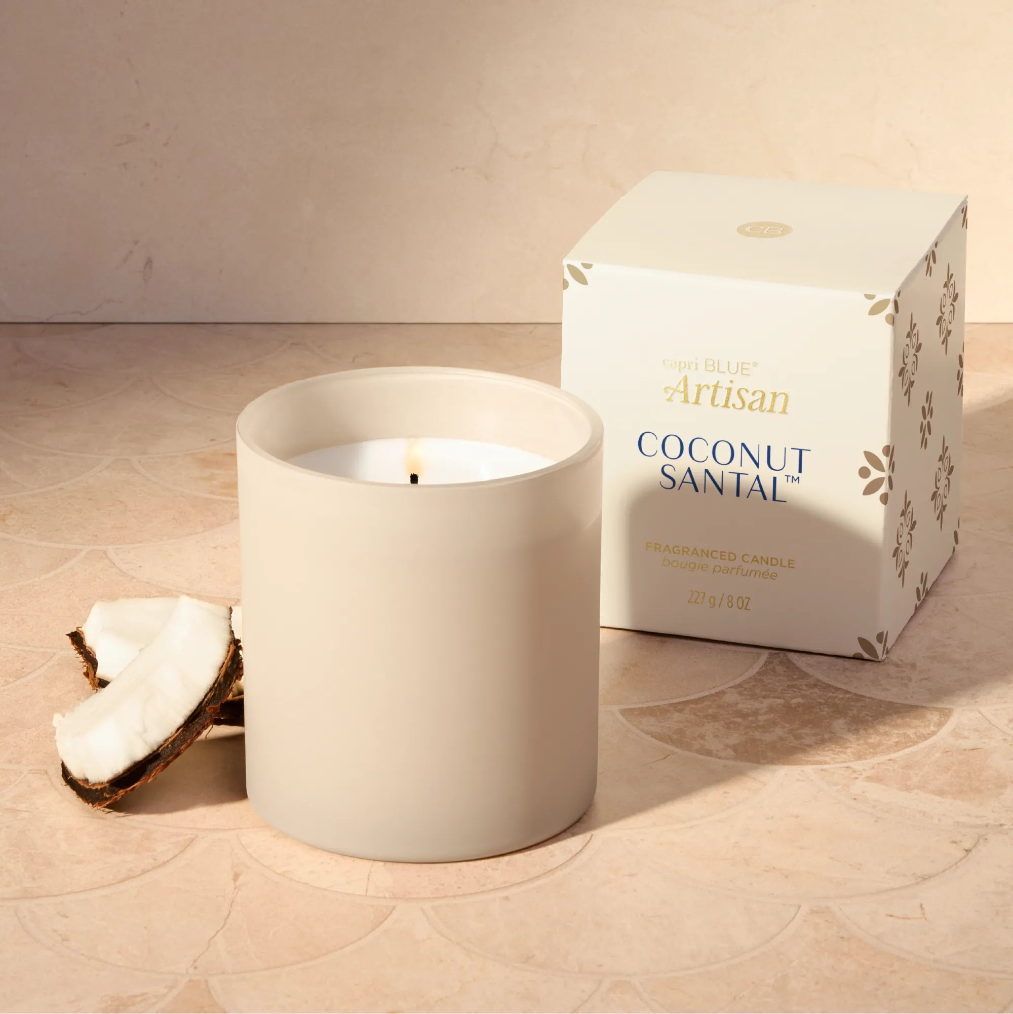 A matte cream-colored soy candle in a matching cylindrical jar sits next to its white packaging box, with a halved coconut prop on a patterned beige surface.