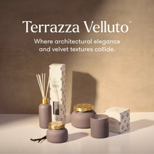 Terrazza Velluto diffuser and candle set in a warm beige studio setting, featuring matte taupe-gray vessels with gold accents, reed sticks, a candle, and branded boxes.