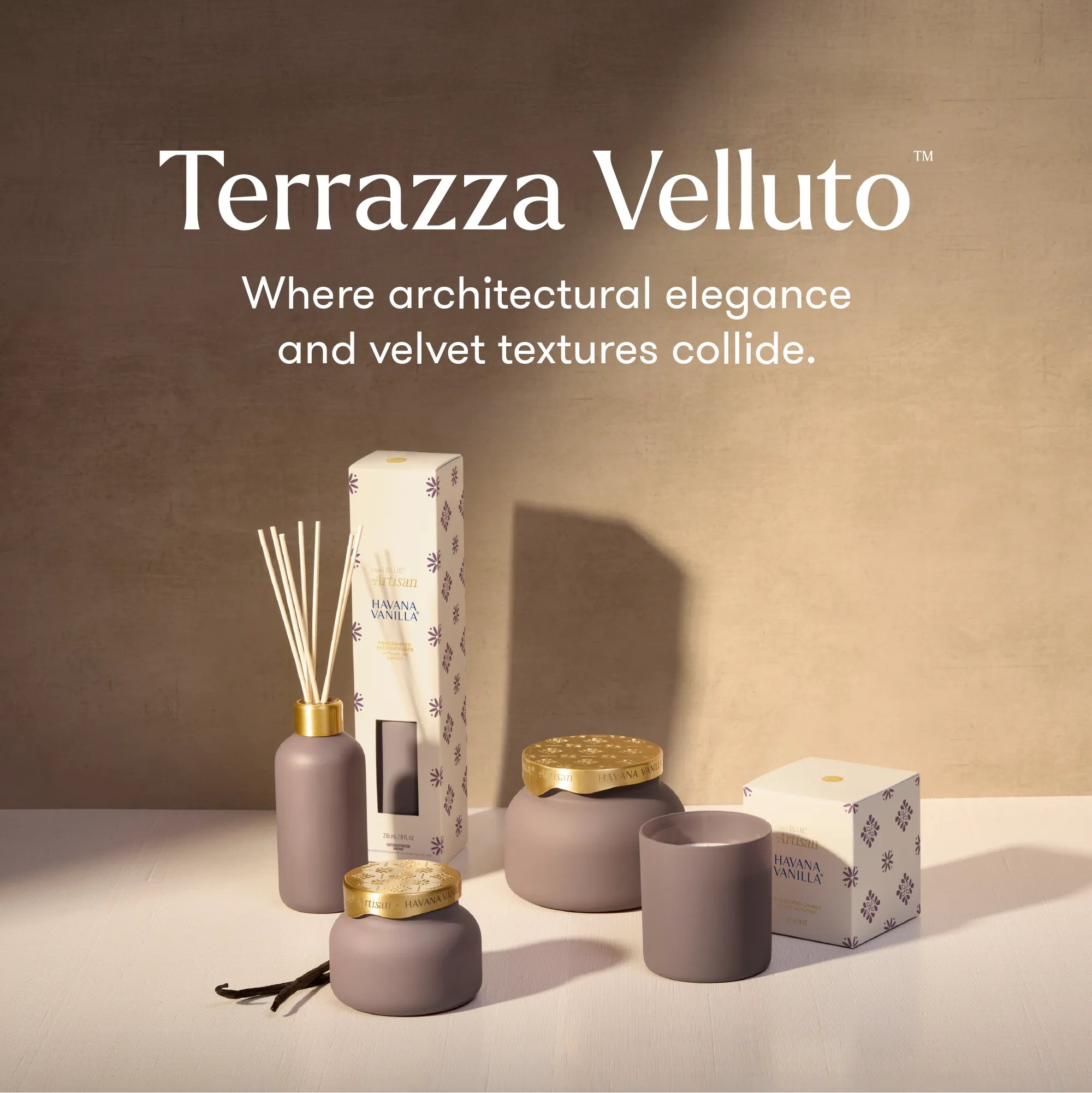 Terrazza Velluto diffuser and candle set in a warm beige studio setting, featuring matte taupe-gray vessels with gold accents, reed sticks, a candle, and branded boxes.