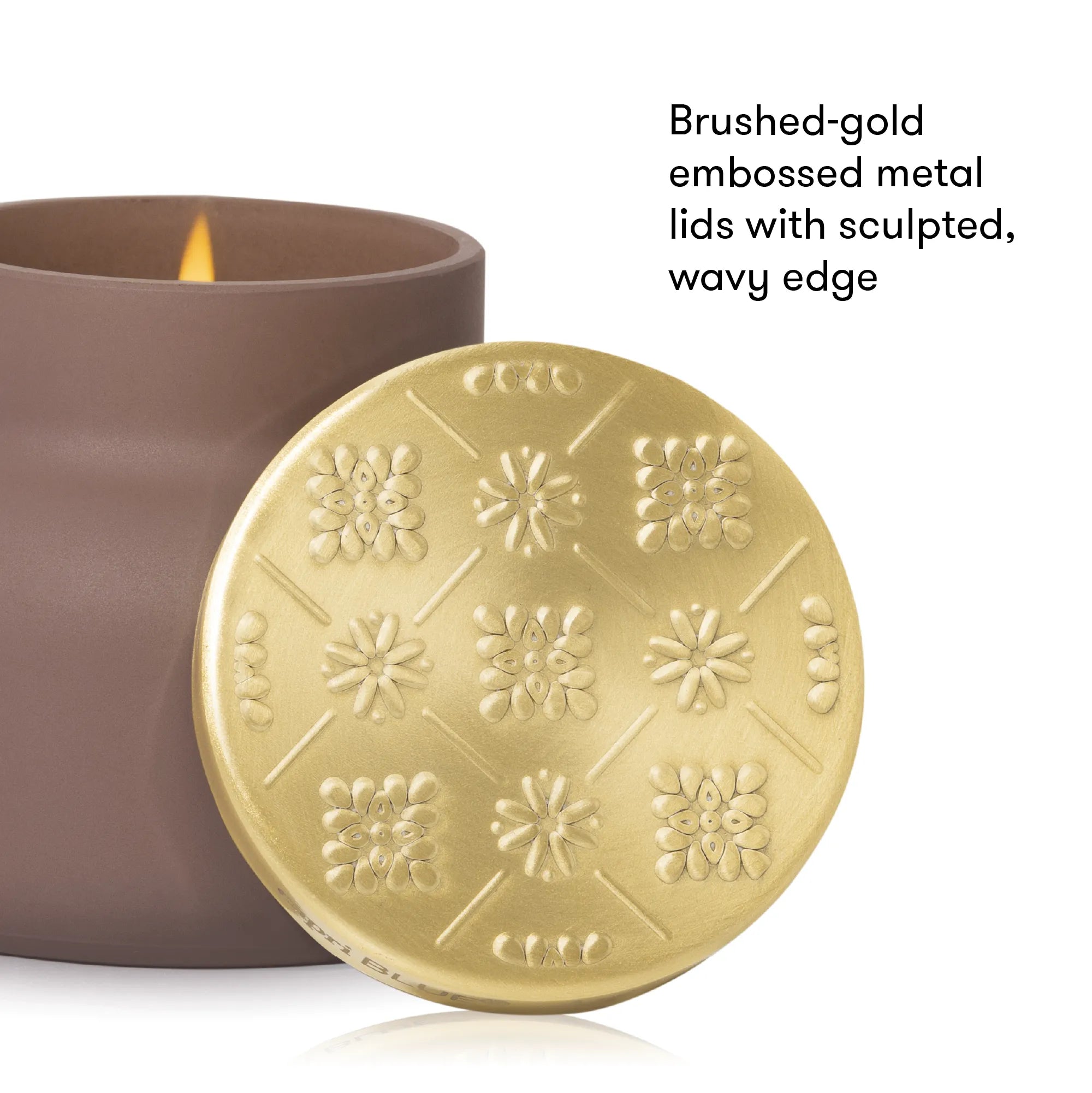 A close-up of a candle jar with a brushed gold embossed metal lid leaning in front, next to the brown candle container. Text on the right reads: 'Brushed-gold embossed metal lids with sculpted, wavy edge.