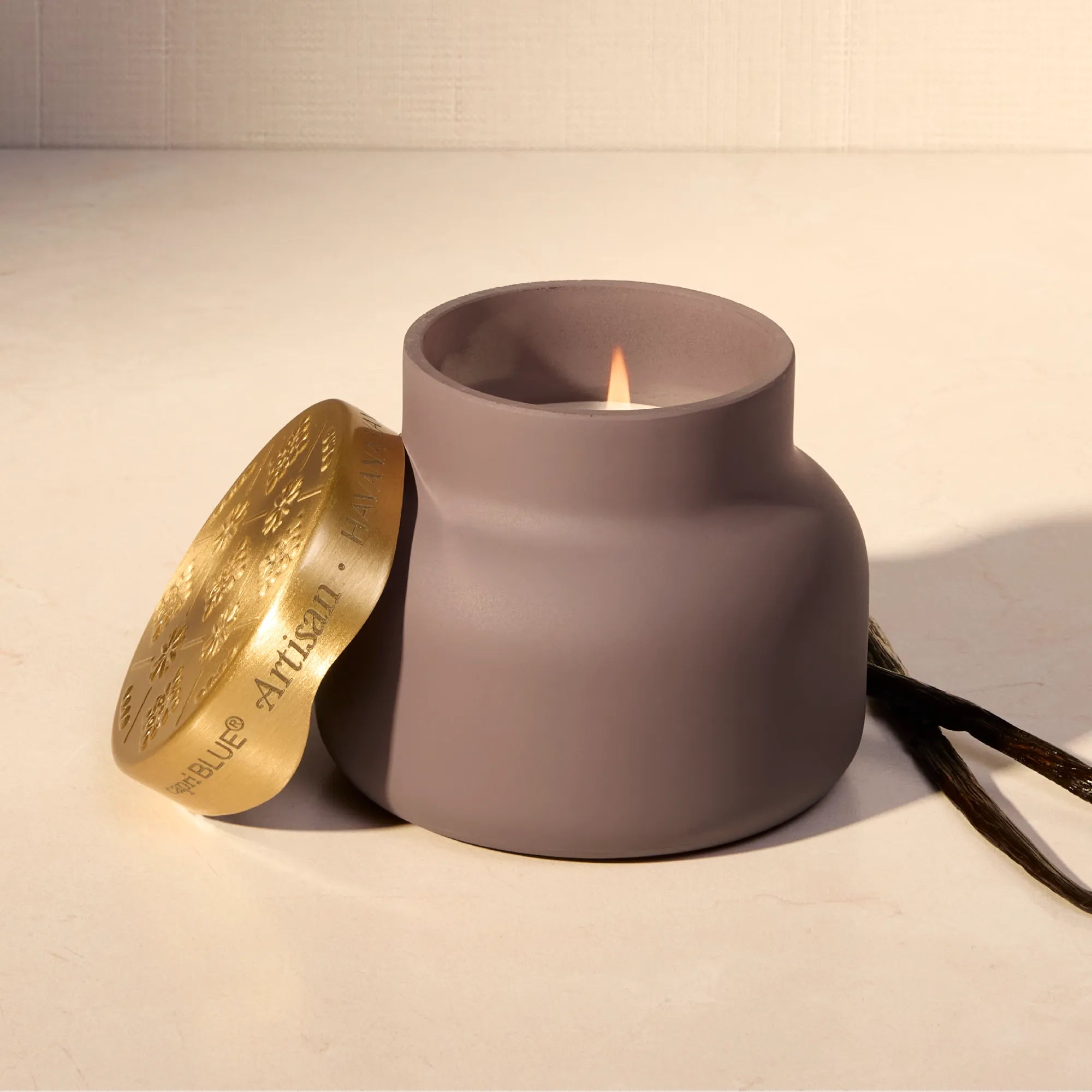 A matte taupe-brown ceramic candle holder with a lit candle inside, shown next to a gold-toned decorative lid engraved with 'Amore Artigiano', on a light beige surface.