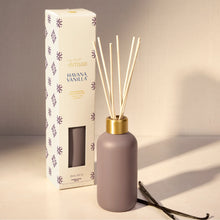 A reed diffuser bottle with natural reeds and a gold cap stands next to its white packaging box with purple floral patterns. The bottle is a muted dusty lavender-gray, and the box is labeled 'Artisan HAVANA VANILLA'.