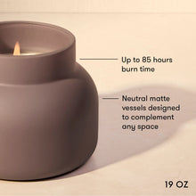 A matte taupe-gray ceramic candle holder with a lit candle, shown with text callouts highlighting its 19 oz size and up to 85-hour burn time on a warm beige background.