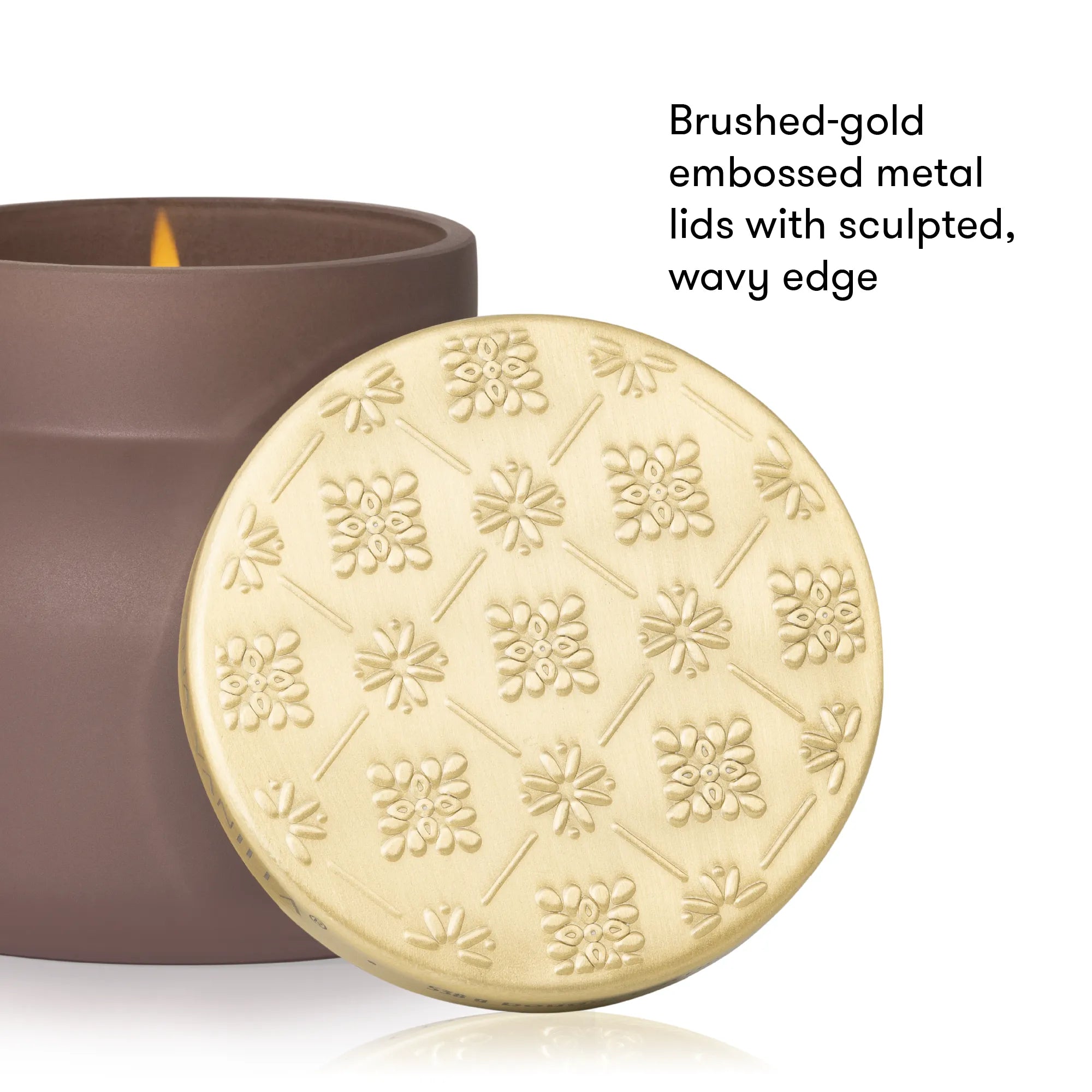 A close-up of a candle jar with its round embossed metal lid leaning in front. The lid is brushed gold with a floral pattern and a wavy edge, and the candle body is dark brown.