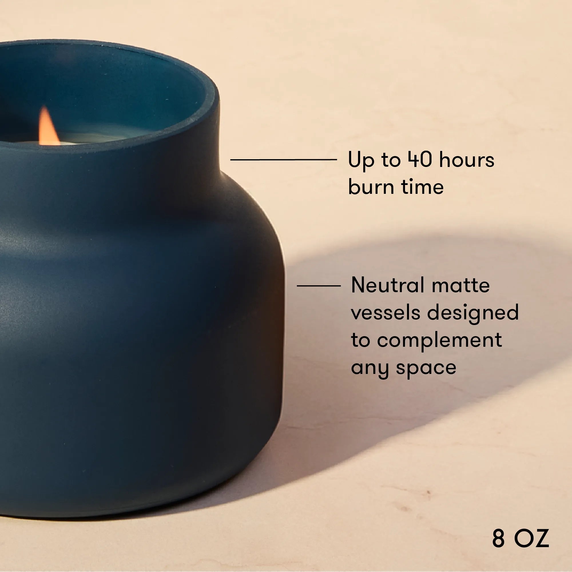 A dark blue matte candle vessel with a lit candle, shown on a beige surface with text overlays describing its 8 oz size and burn time.