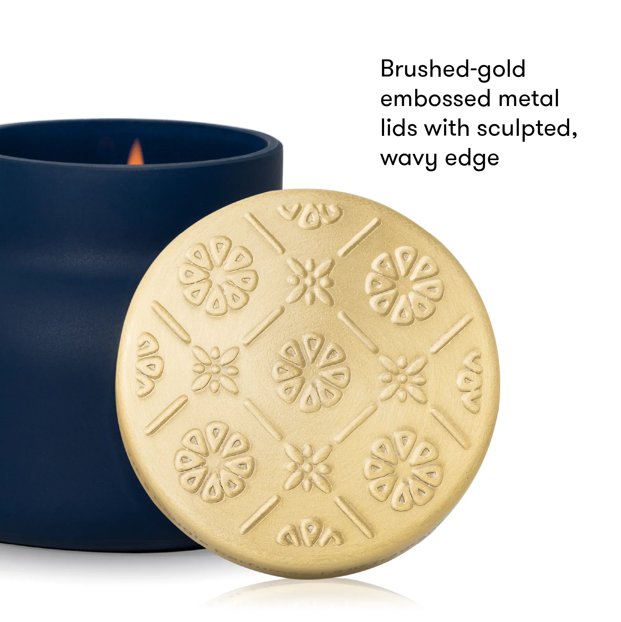 A close-up of a round brushed-gold metal candle lid with embossed floral and geometric patterns, shown in front of a dark navy blue candle container with a visible flame. Text on the right reads, 'Brushed-gold embossed metal lids with sculpted, wavy edge.