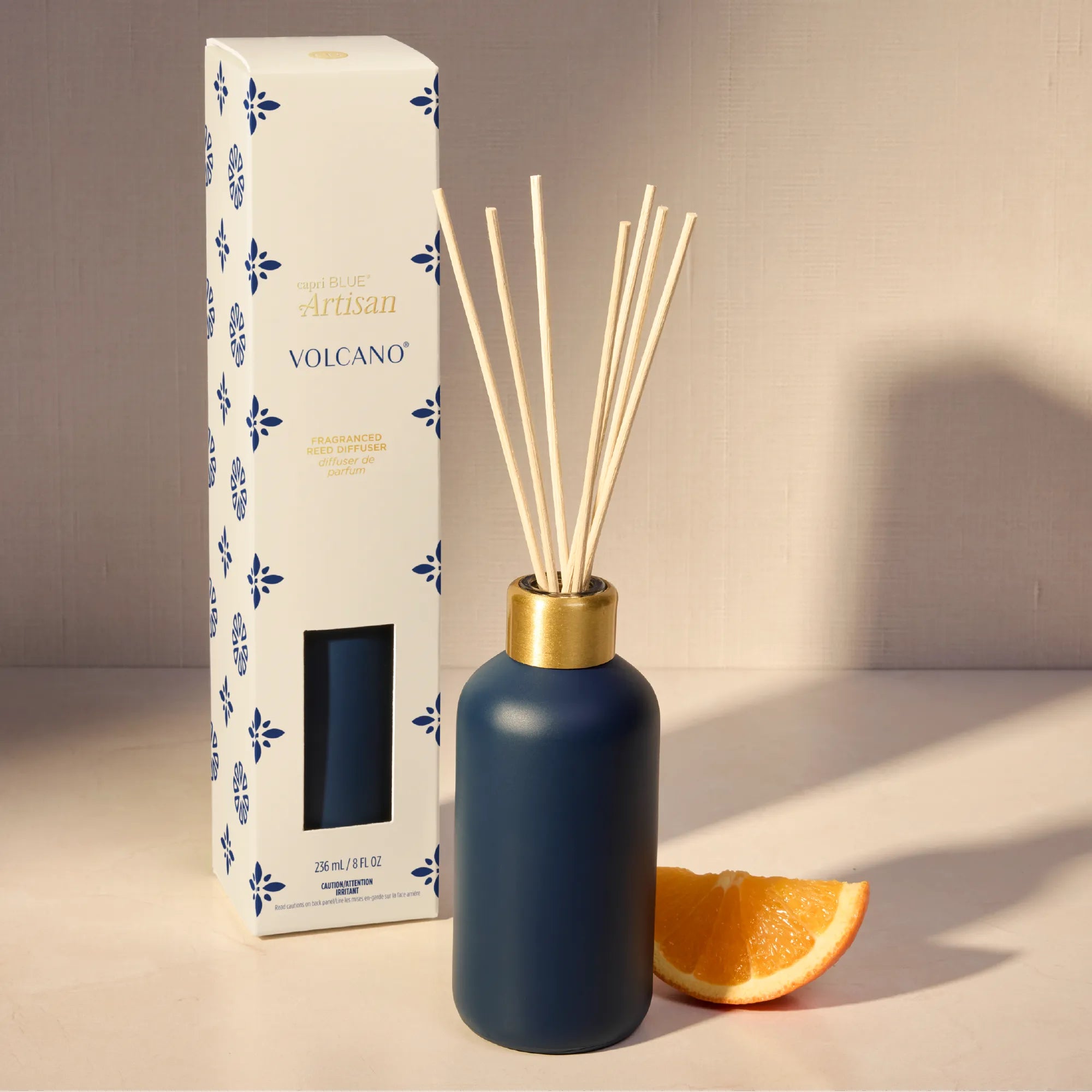 A reed diffuser bottle with a matte navy blue body, a gold collar, and natural reeds, shown next to its white and blue patterned packaging box and a slice of orange on a beige surface.
