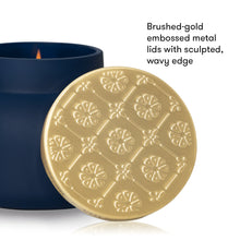 A brushed gold metal lid with an embossed floral pattern and wavy edge, shown leaning against a dark navy blue candle jar on a white background. Black text on the right reads: 'Brushed-gold embossed metal lids with sculpted, wavy edge.