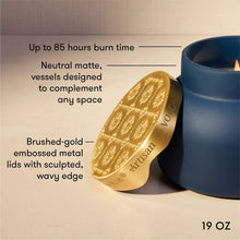 A 19 oz scented candle wax melt by Artisan Volatility, shown with its brushed gold metal lid leaning against a blue ceramic container. The image includes text callouts highlighting features like up to 85 hours of burn time, neutral matte vessels, and the brushed-gold embossed lid.