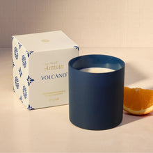 A navy blue glass jar candle with white wax sits next to its white and blue patterned packaging box, with a slice of orange on a beige surface.
