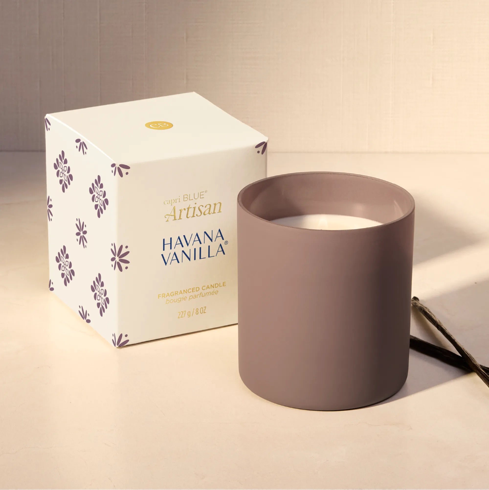 A matte taupe-gray cylindrical candle jar next to its white and purple patterned retail box, with a lit match on a warm beige surface.