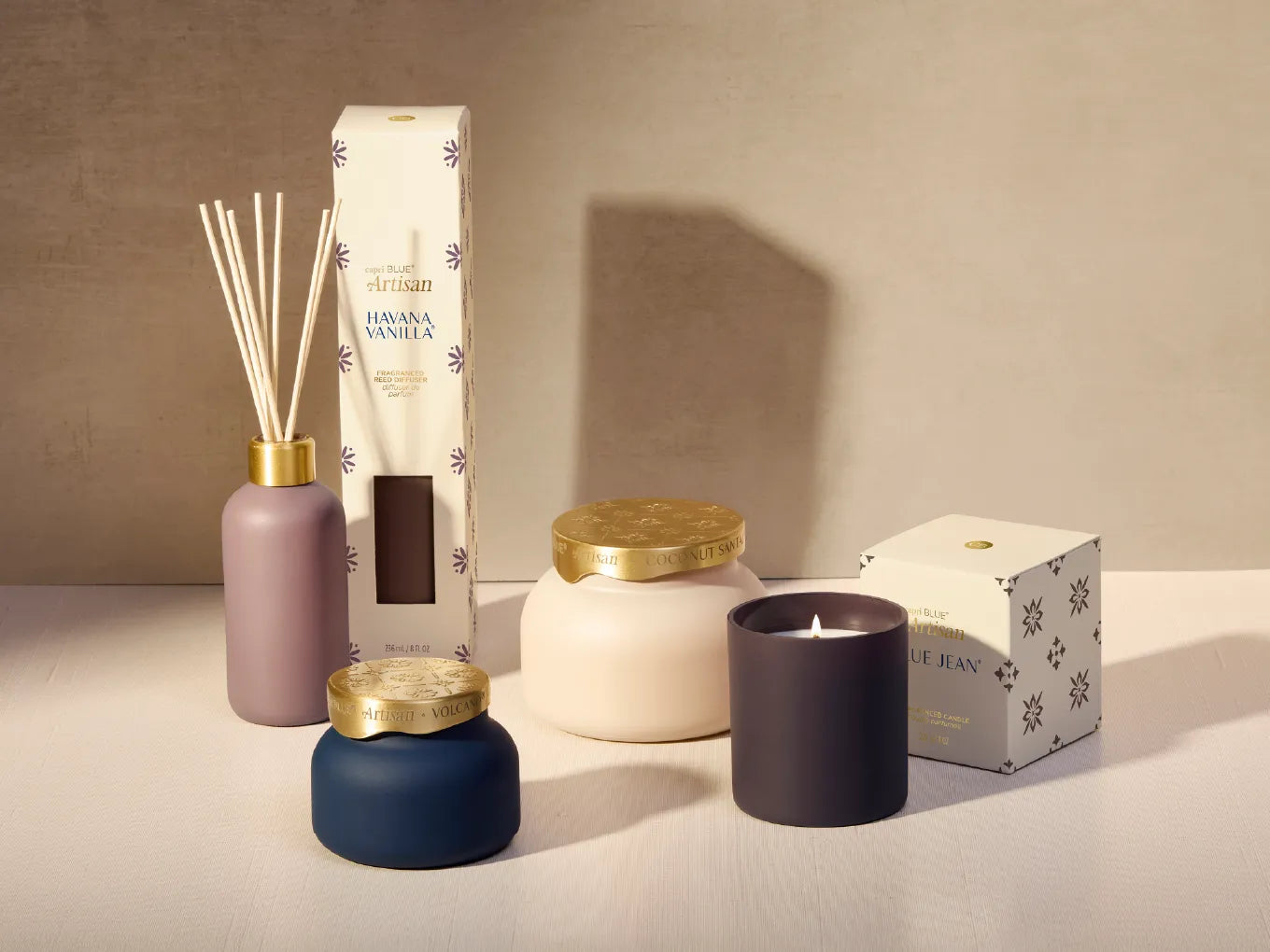 A curated collection of home fragrance products including a reed diffuser, a jar candle, a scented candle, and packaging boxes, styled on a neutral beige surface against a matching background.